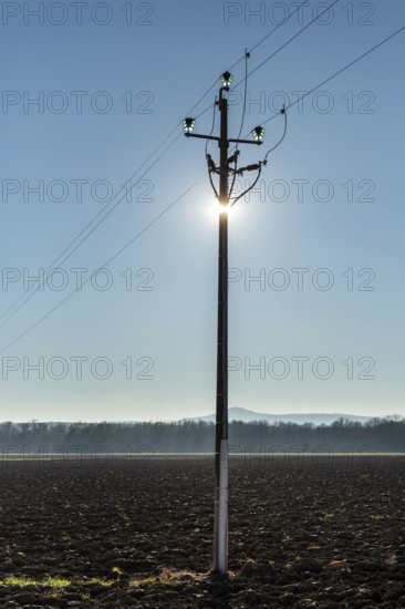 A power pole stands tall in the foreground, with sunlight shining directly behind it, creating a glowing silhouette. The background features an expansive field under a blue sky. France