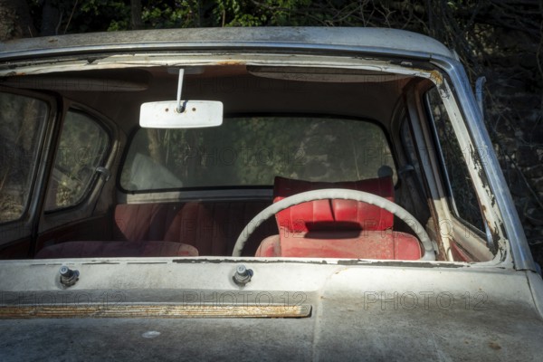 A deserted vintage car sits in a state of disrepair, showcasing a cracked windshield and a faded interior. The red seat adds a pop of color under soft, natural light