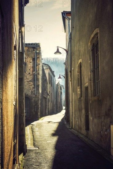 A narrow cobblestone street in an ancient village reflects the soft morning light and shadow. The warm glow illuminates weathered buildings lining the path. France