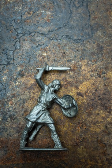 A detailed warrior figure stands ready for battle, gripping a sword in one hand and a shield in the other. Its metallic texture contrasts with the earthy backdrop, suggesting a fierce engagement