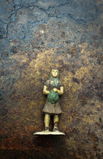 A small, weathered figurine depicting a girl in a dress, clutching a green bundle, stands on a rough, textured surface, showing the passage of time with its aged colors