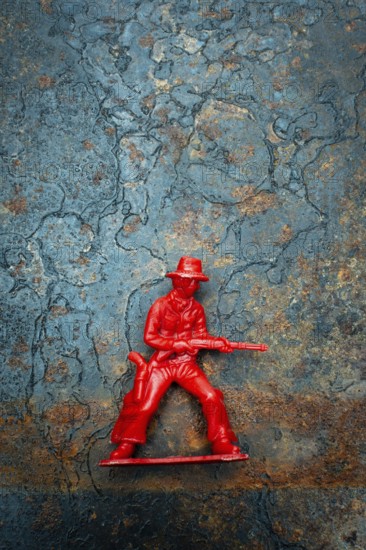 A small red plastic toy cowboy stands firmly on a rustic blue and brown textured background. The figure is equipped with a weapon