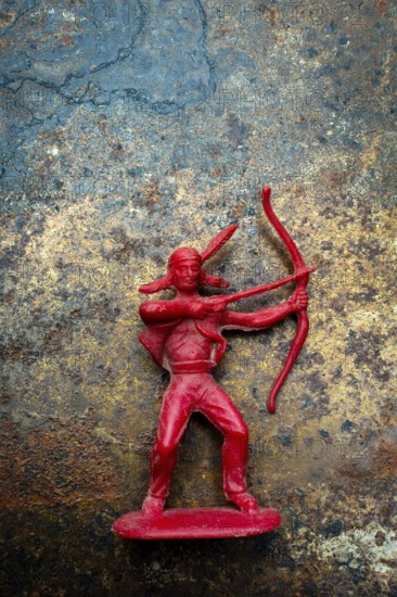A red plastic toy archer represents a Native American figure, posed with a bow and arrow on a textured, rust-colored surface. T