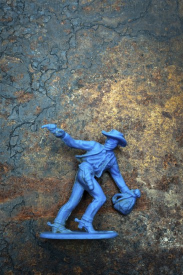 A bright blue cowboy figure stands firmly on a rugged surface, one hand raised with a pistol while the other holds a bag. The contrast highlights its playful details against a worn backdrop