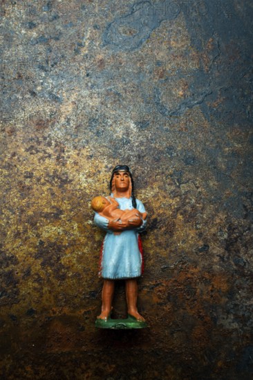 A vibrant plastic figure represents an Indian Native American woman cradling a child, positioned on a rustic, weathered surface