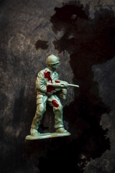 A green soldier figurine holds an assault weapon while displaying battle wounds marked by red stains. The dark backdrop serves to enhance the gravity of war and its emotional toll