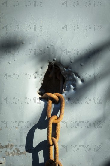 A rusty chain is situated through a weathered hole in a blue surface, with sunlight creating shadows that highlight the textures and condition of the surrounding materials