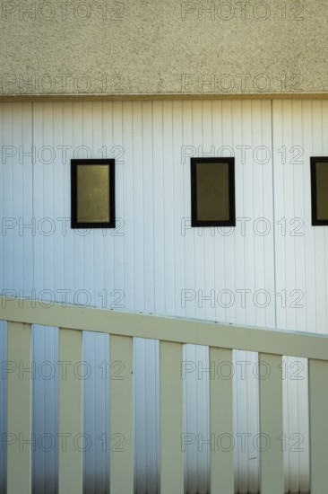 A building featuring three rectangular windows on a white vertical paneled wall. A white railing leads up to the structure, hinting at a modern architectural style during sunset
