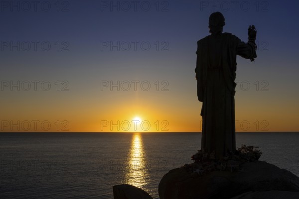 The sun rises on the Costa Rei, a stretch of coastline on the Italian Mediterranean island of Sardinia. In front of it is the silhouette of a 1.80 metre high bronze statue of Christ called the Blessing Christ of Santa Giusta, also known as Christ the Navigator, Monte Nai, Costa Rei, Sardinia, Italy