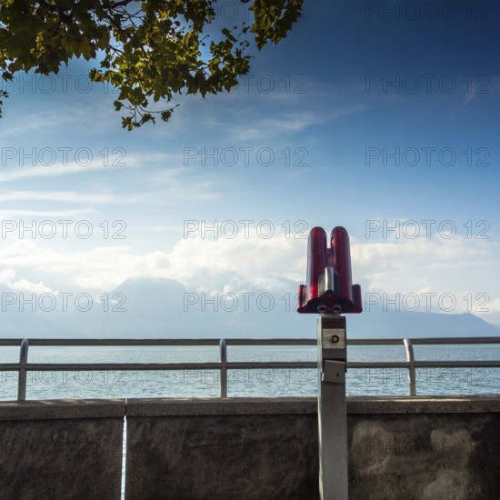 A telescope stands on the lakeside walkway in Vevey, Switzerland, overlooking Lake Genova. The clear sky and distant mountains create a picturesque backdrop for visitors. Switezerland