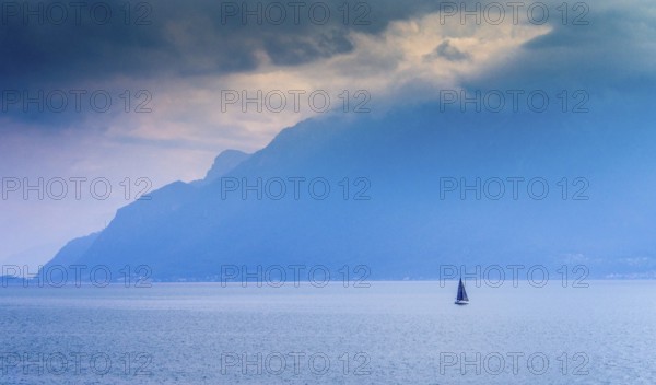 A lone sailboat navigates the tranquil waters of Lake Geneva surrounded by shadowy mountains. The atmosphere is calm yet dramatic under the cloudy sky. Switzerland