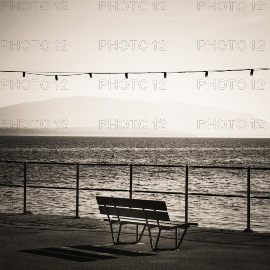 A solitary bench sits by the lakeside promenade in Nyon, Switzerland. Gentle waves ripple in Geneva Lake while distant mountains create a tranquil backdrop, enhancing the peaceful atmosphere