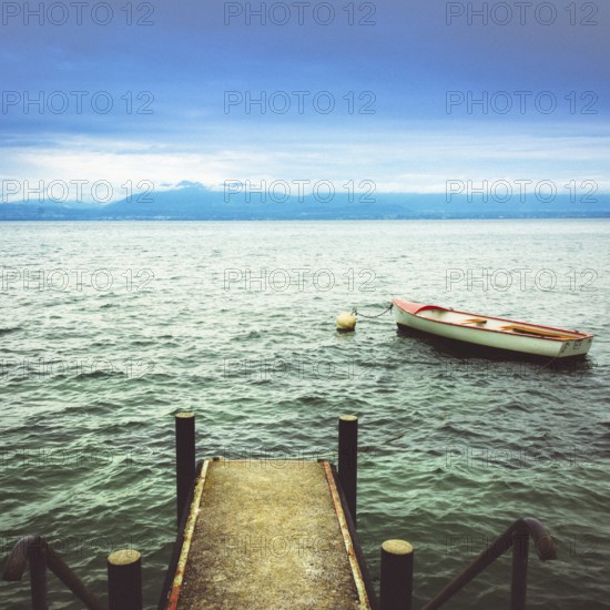 A wooden jetty reaches into the calm waters of Lake Geneva, where a small boat is moored. Mountains outline the horizon under a cloudy sky, showcasing the tranquil beauty of Switzerland