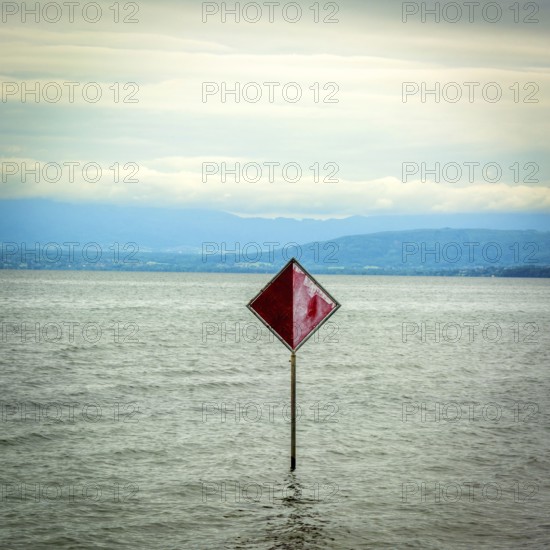 A prominent red information sign rises above the water of Lake Geneva, indicating safety or navigation. The scenery features a serene lake with distant hills visible against a cloudy sky. Switzerland