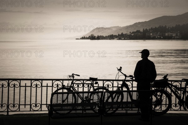 A tourist stands at the edge of Lake Geneva, captivated by the serene waters and distant hills. Bicycles are parked nearby, creating a peaceful atmosphere for exploration at sunrise. Switzerland