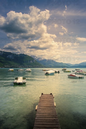 A wooden pontoon extends over the tranquil waters of Annecy Lake, surrounded by mountains and dotted with colorful boats. The sky is partially covered by clouds, creating a serene atmosphere. Haute Savoie. Auvergne Rhone Alpes. France