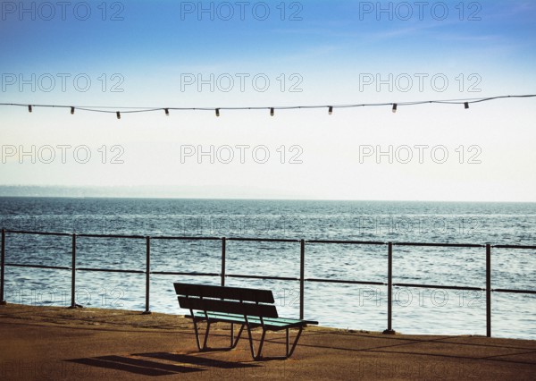 A bench is positioned along the promenade beside Geneva Lake in Nyon, Switzerland, providing a peaceful spot to enjoy stunning views of the calm water and clear blue sky