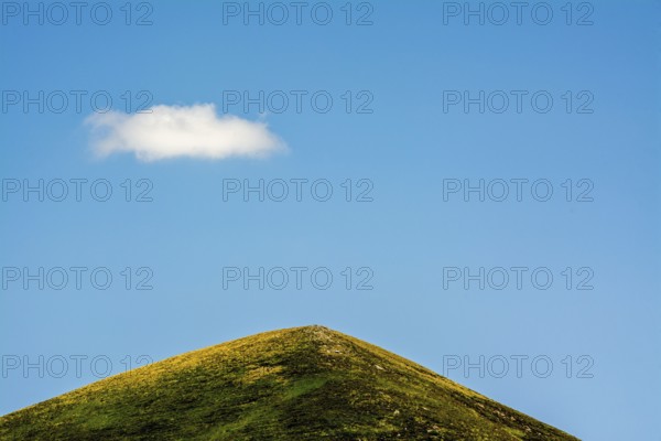 A solitary cloud floats above a green mountain in the Auvergne-Rhone-Alpes region of France. The landscape is tranquil, showcasing nature's beauty under a vast blue sky