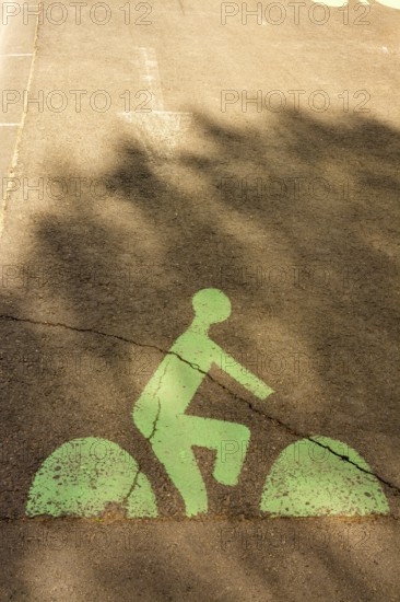 A green bicycle lane marking is visible on a city street, featuring a cyclist silhouette. Shadows from trees create a pattern over the pavement, adding depth to the scene