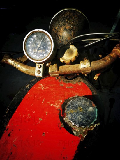 A worn motorcycle dashboard features a rusty speedometer and a faded paint job. The dark background emphasizes the aged components, highlighting the passage of time