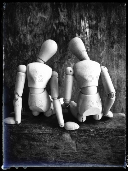 Two wooden figures are positioned closely together, facing away from the viewer. They sit on a rustic wooden surface, showcasing their joint form in a serene and artistic manner