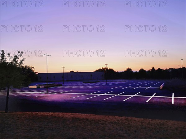 A quiet parking lot in France at dusk, illuminated by streetlights. The clear sky transitions from deep blue to vibrant orange, casting an inviting glow over the empty spaces. France