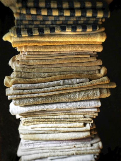 A neat stack of folded textiles in various shades of beige, cream, and a hint of black is illuminated by soft, natural light, enhancing their textures and warmth