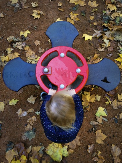 A young child is playing with a vibrant red spinner at a playground, surrounded by fallen yellow and brown leaves, capturing the essence of autumn joy and exploration