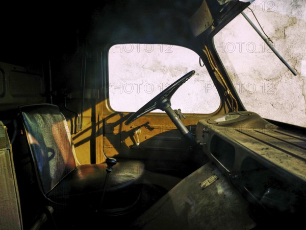 An abandoned truck interior features worn seats and an aged steering wheel, casting shadows against the bright wall. The play of light highlights the passage of time