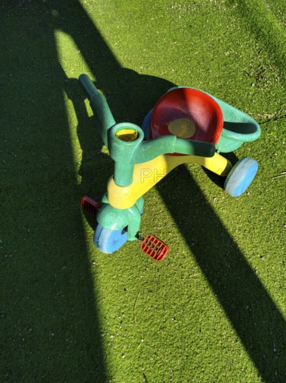 A vibrant, multi-colored tricycle sits on lush, green artificial grass, casting a shadow in bright sunlight. Its playful design invites children's imagination and outdoor fun