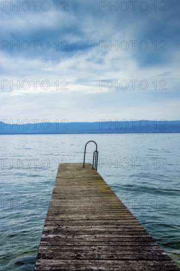 A sturdy jetty extends into the serene waters of Lake Geneva, surrounded by mesmerizing blue hues and a tranquil atmosphere. Perfectly captures the beauty of Haute-Savoie on a peaceful day, Auvergne Rhone Alpes, France