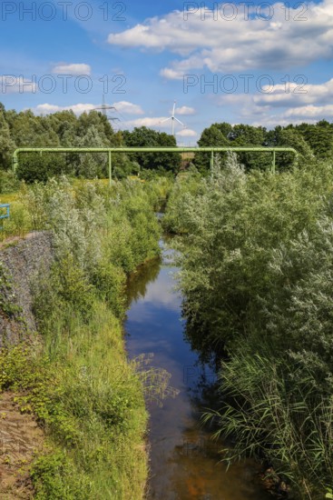 Bottrop, North Rhine-Westphalia, Germany - Renaturalised Boye, the tributary of the Emscher, has been transformed into a near-natural watercourse, flood protection through newly designed flooding areas, the Boye is now free of wastewater following the construction of a parallel sewer, is part of the Emscher river system and thus part of the Emscher conversion, was previously an open, above-ground wastewater channel, mixed water channel with surface water and wastewater. Wind turbine on the Mottbruch spoil tip at the back