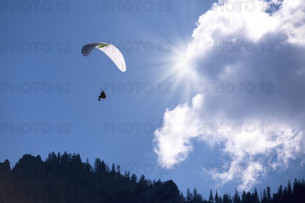 A paraglider flies under a bright sun and clouds in the blue sky, Bavaria, Germany