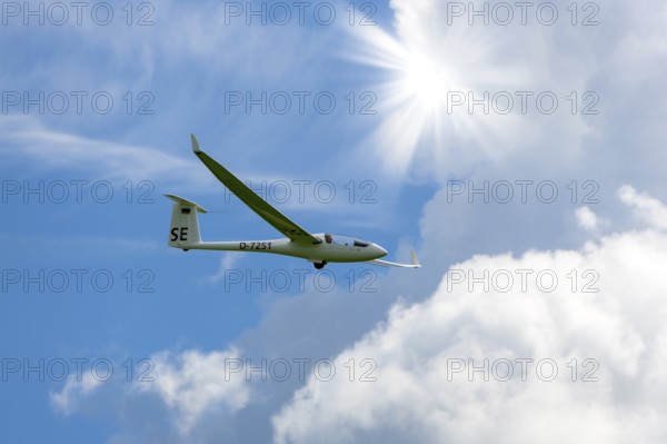 A glider flies through a blue sky with white clouds and bright sun, Baden-Württemberg, Germany