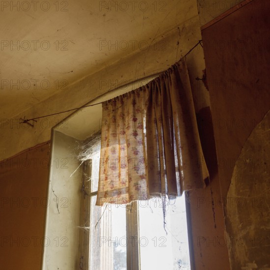 Sunlight streams through a dusty window adorned with worn curtains, highlighting the cobwebs in a neglected room. Dust particles float in the air, evoking a sense of nostalgia