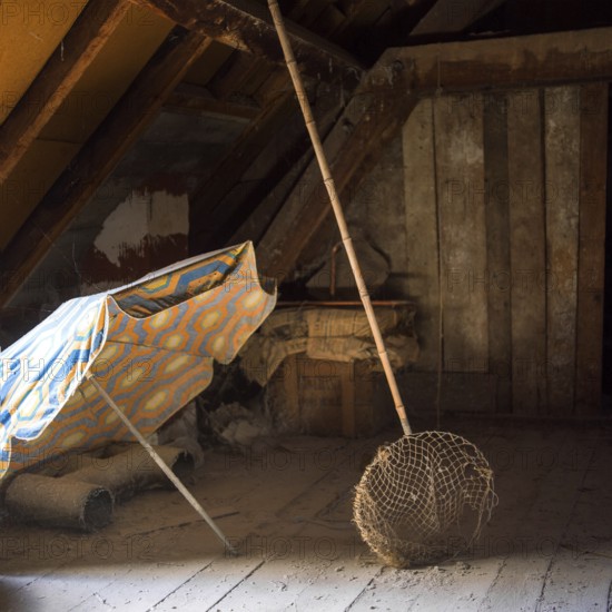 Sunlight filters through an attic, illuminating a colorful sunshade and a butterfly net alongside dust-covered wooden beams, creating a tranquil, nostalgic atmosphere