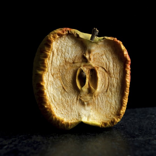 A half apple displays its dried, wrinkled surface with unique textures and patterns. The apple sits on a dark stone surface, highlighting its aged appearance. The light emphasizes its natural tones