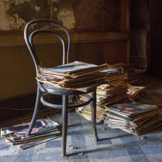 An old chair is perched beside a towering stack of newspapers, both covered in dust, in a dimly lit room that suggests neglect and the passage of time, evoking memories of a bygone era