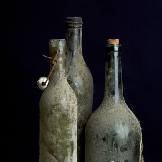 Three dusty vintage bottles stand together, covered in grime, highlighting their age and history. These relics evoke memories of past gatherings and forgotten stories in a dimly lit cellar