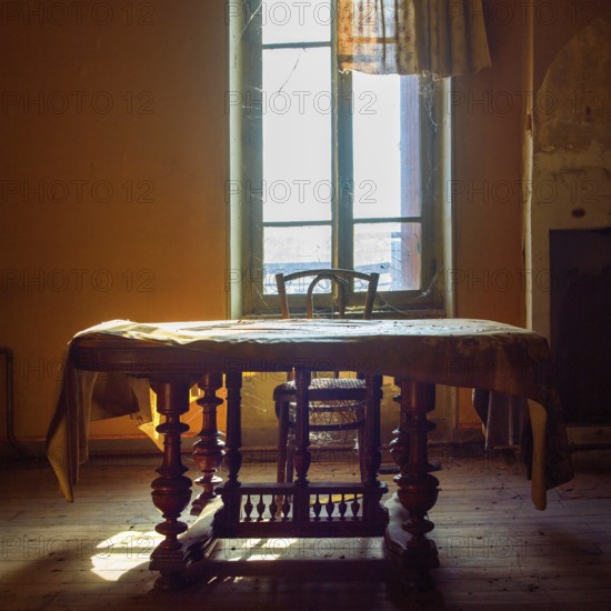 Warm sunlight filters through an old window, illuminating a vintage wooden table and chair. Dust gathers in the quiet room, evoking a sense of nostalgia and forgotten stories