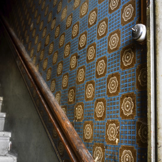 A close-up view of old wallpaper lining the walls of a staircase corridor, featuring a geometric pattern with floral motifs and a wooden handrail that adds a rustic touch