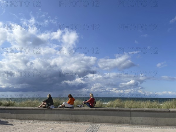 Three holidaymakers enjoying the sun, sitting on a little wall on the promenade, blue sky, Baltic resort Kühlungsborn, Baltic Sea, Mecklenburg-Western Pomerania, Germany
