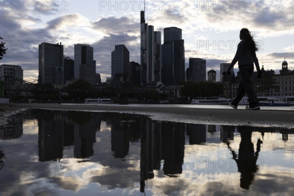 A woman walks along the banks of the Main in the rain in front of the Frankfurt banking skyline, Frankfurt am Main, Hesse, Germany