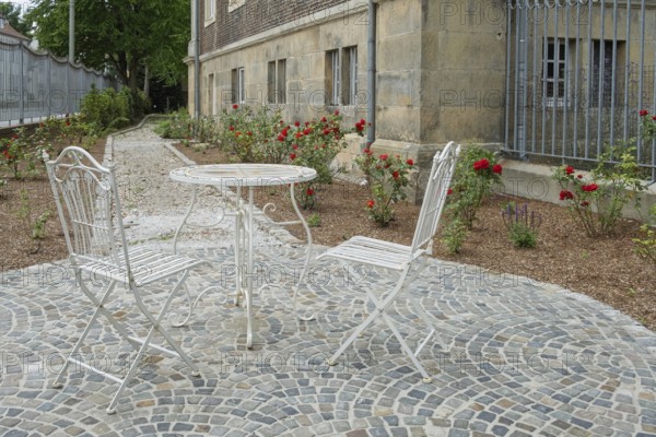 Two white metal chairs and a table on a paved square in front of a historic building, surrounded by roses, Münsterland, North Rhine-Westphalia, Germany