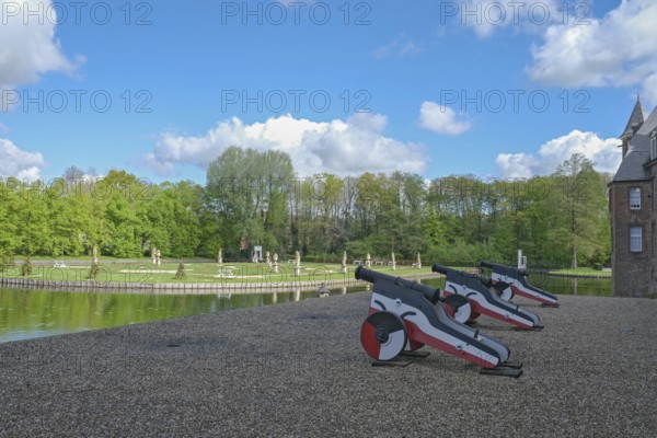 Small cannons at the edge of a park with a pond and sculptures, under a blue sky with clouds, moated castle Anholt, Münsterland, North Rhine-Westphalia, Germany