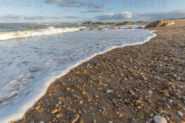 A wide sandy beach stretches out with scattered pebbles and beach shells. Gentle waves crash against the shore under a partially cloudy sky during late afternoon. Plage de l'Aubraie, Les Sables d'Olonne, Vendee, France