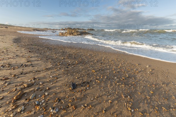 The waves crash gently against a rocky shoreline at sunset, creating a tranquil atmosphere. The sandy beach is strewn with pebbles and surrounded by a beautiful sky. Plage de l'Aubraie, Les Sables d'Olonne, Vendee, France
