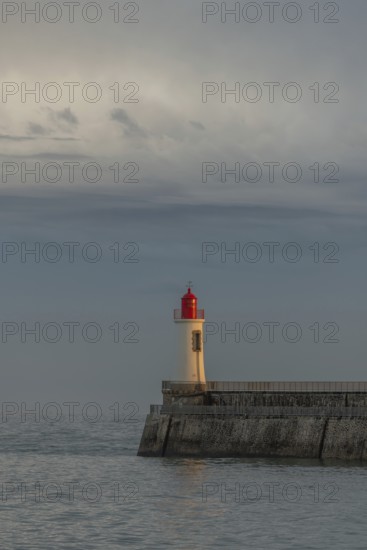 A lighthouse stands on a pier and casts its light over calm waters at dusk. The sky is filled with dramatic clouds, adding to the tranquil atmosphere of the coastal landscape. Les Sables d'Olonne, Vendee, France