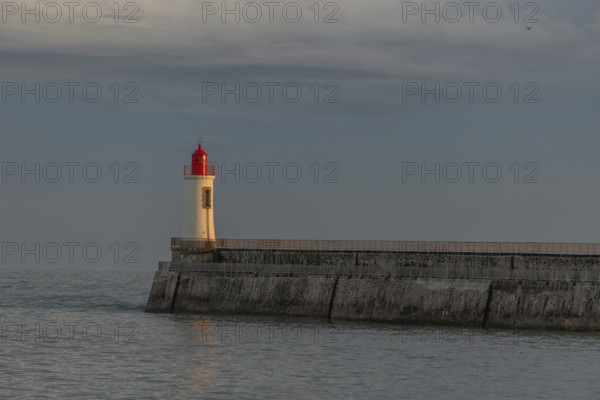 A lighthouse stands on a pier and casts its light over calm waters at dusk. The sky is filled with dramatic clouds, adding to the tranquil atmosphere of the coastal landscape. Les Sables d'Olonne, Vendee, France