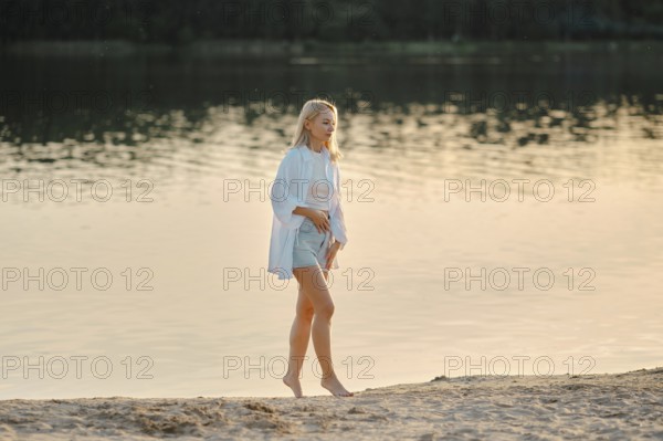 A woman strolls barefoot on a sandy beach by the lake, enjoying the serene atmosphere as the golden hour casts a warm glow over the water and landscape. The soft light enhances the peaceful scenery